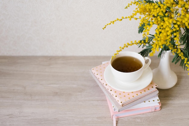 Um buquê de mimosa em um vaso branco uma xícara de chá em uma pilha de cadernos para anotações