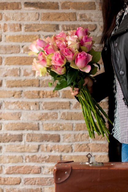 Um buquê de rosas nas mãos de uma mulher com uma mala perto de uma parede de tijolos