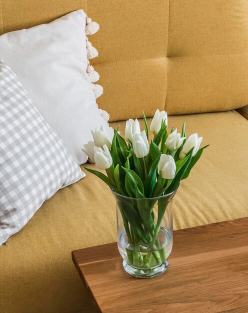 Um buquê de tulipas brancas em um vaso de vidro em uma mesa de madeira ao lado de uma poltrona