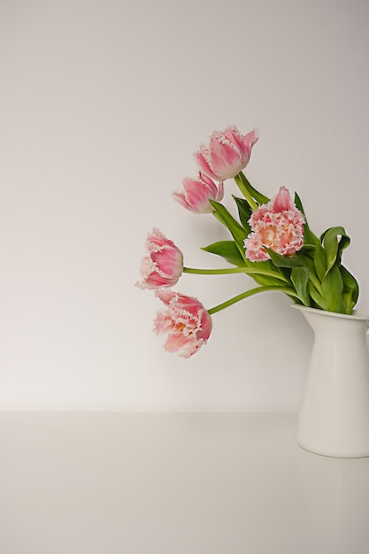 Um buquê de tulipas papagaio rosa fresco com folhas verdes em um jarro de cerâmica branca de pé sobre uma mesa contra o pano de fundo de papel. Composição decorativa da flor.