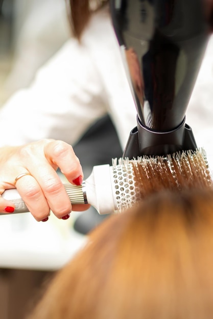 Um cabeleireiro profissional está secando longos cabelos ruivos com secador de cabelo e escova redonda, close-up.