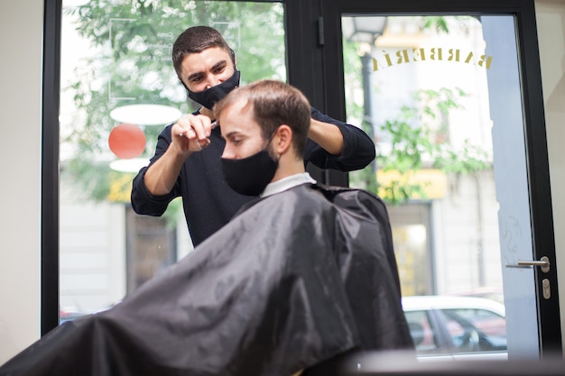 Um cabeleireiro profissional usando máscara protetora cortando o cabelo de uma cliente durante o coronavírus