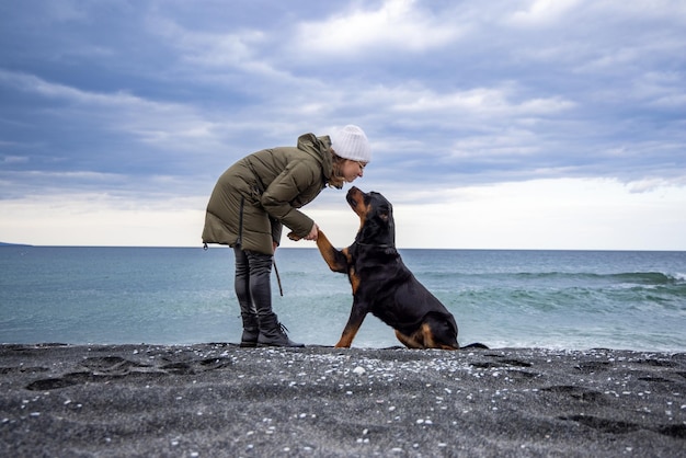 Um cachorro em uma coleira senta-se na praia e dá uma pata a uma mulher em clima frio