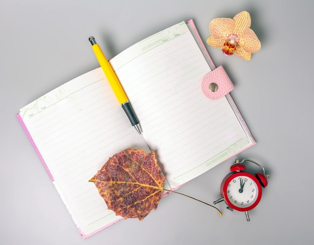 Um caderno aberto, caneta, flor de orquídea e um despertador que lembra o tempo estão sobre a mesa.