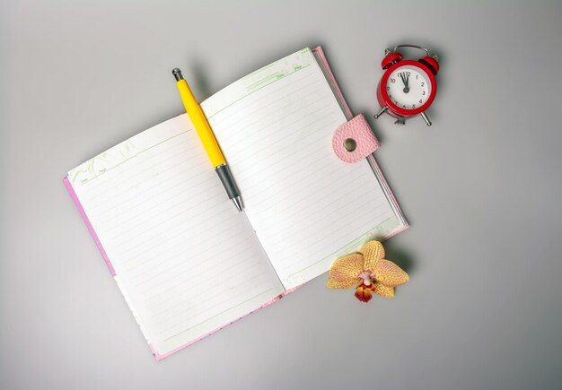 Um caderno aberto, caneta, flor de orquídea e um despertador que lembra o tempo estão sobre a mesa.