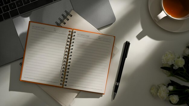 Um caderno aberto com páginas em branco e material de escritório na mesa sob a vista superior da sombra da luz solar