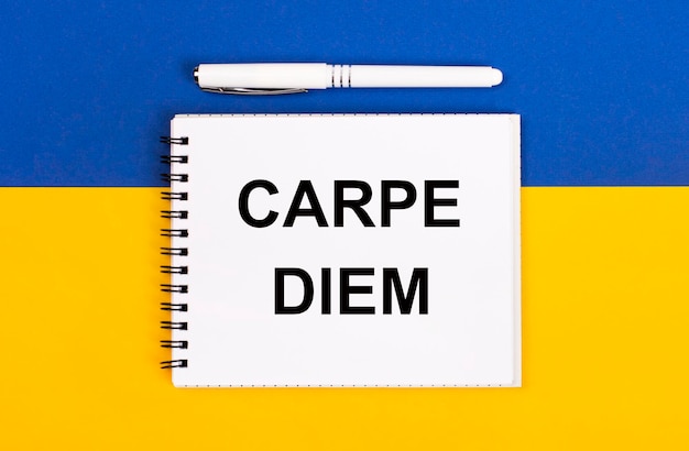 Um caderno branco com o texto CARPE DIEM e uma caneta branca sobre fundo azul e amarelo