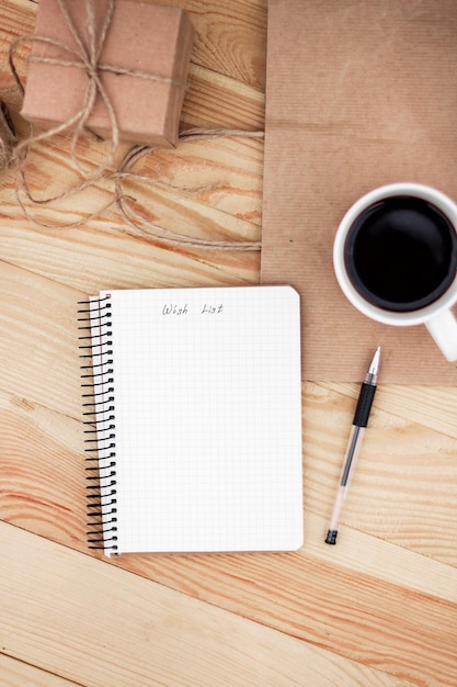 Um caderno com uma lista de desejos de inscrição deitado na mesa de madeira. Está rodeado por caneta preta, chávena de café, presente e saco de papel.