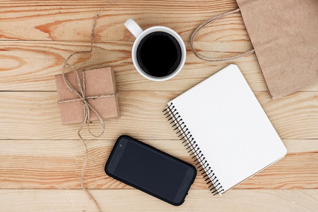 Um caderno em branco deitado na mesa de madeira. Está rodeado de presente, smartphone, chávena de café, caneta preta e saco de papel.