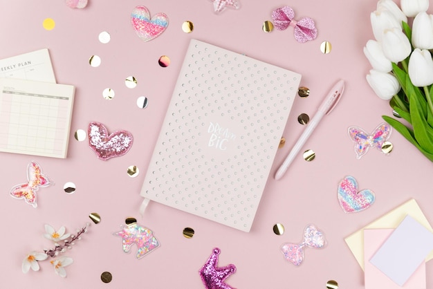 Um caderno rosa com uma caneta rosa em um fundo rosa com confetes e corações.