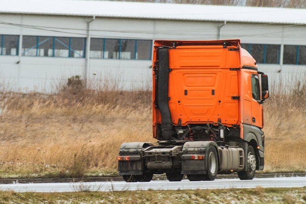 Um camião viaja por uma estrada aberta sem reboque. Um camião moderno europeu funciona com motor diesel. A estrada é flanqueada por grama selvagem e uma área industrial à distância.