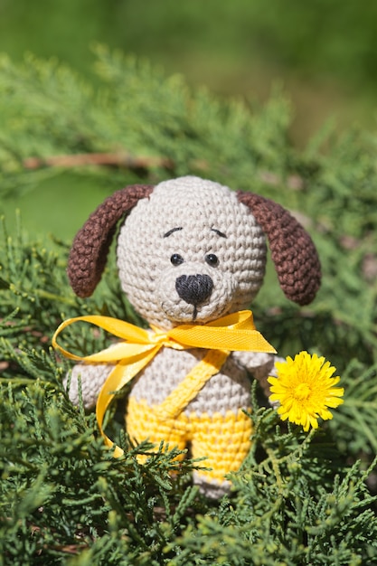 Um cão marrom feito malha pequeno com uma fita amarela no jardim do verão. Brinquedo de malha, feito à mão, amigurumi