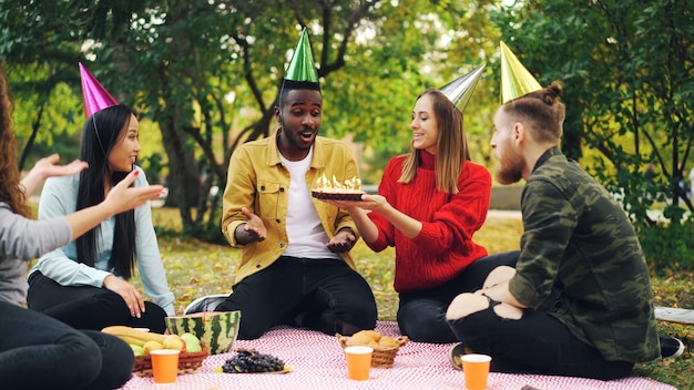 Um cara afro-americano barbudo está fazendo uma festa de aniversário no parque soprando velas no bolo e rindo desfrutando de surpresa que seus amigos estão batendo palmas