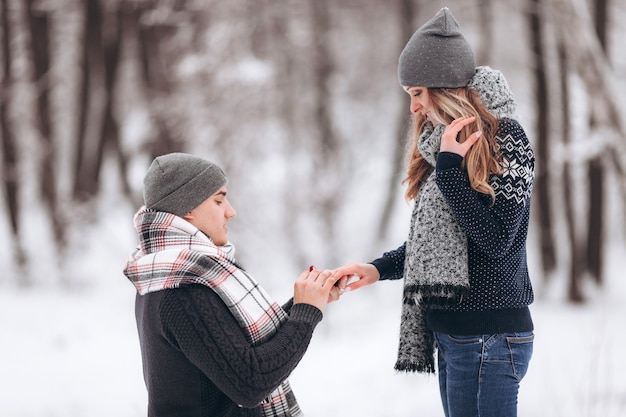 Um cara ajoelhado coloca uma aliança de casamento na mão de uma garota, fazendo uma proposta de casamento em um bosque nevado no inverno