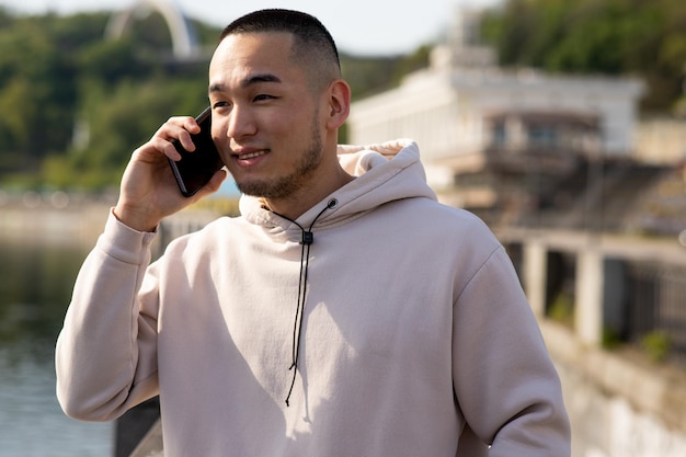 Um cara asiático feliz com capuz esportivo está falando ao telefone com um sorriso no fundo desfocado da natureza