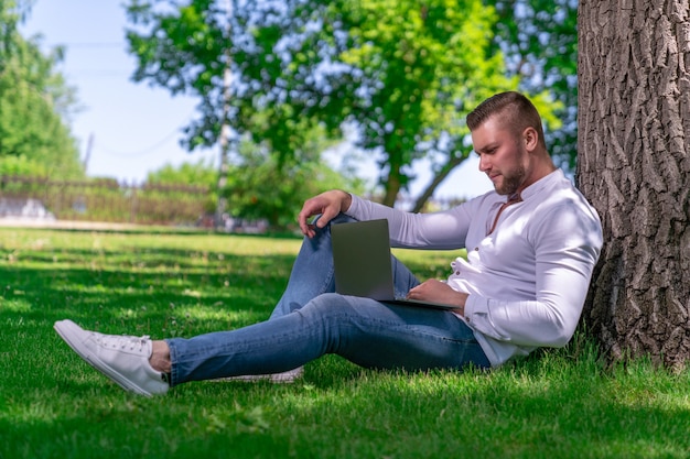 Um cara bonito com cabelo loiro e barba por fazer está sentado na grama perto de uma árvore em um parque rural com um laptop aberto no colo