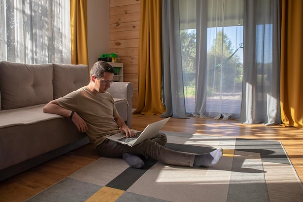 Um cara com roupas casuais está sentado com um computador e trabalhando no chão em um apartamento ensolarado