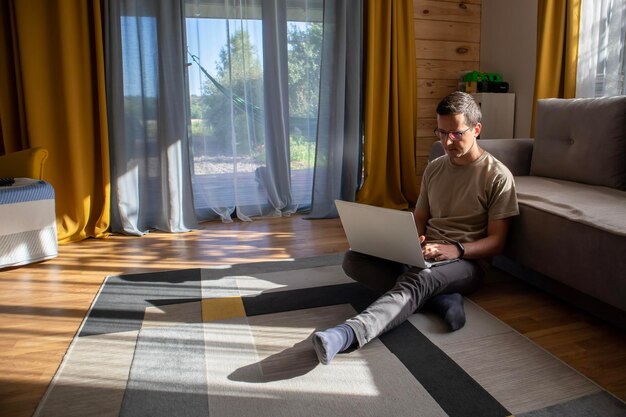 Um cara com roupas casuais está sentado com um computador e trabalhando no chão em um apartamento ensolarado