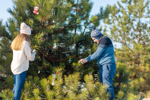 Um cara com uma garota decora uma árvore de Natal verde em uma rua no inverno na floresta com brinquedos decorativos e guirlandas. Enfeites de natal