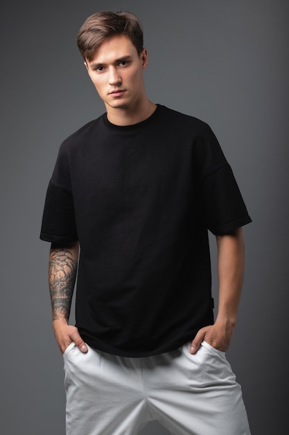 Um cara de aparência de modelo em roupas pretas posa em um fundo cinza de estúdio. Retrato de moda masculina