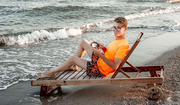 Um cara de camiseta e shorts segura um tablet, sentado em uma espreguiçadeira de madeira à beira-mar