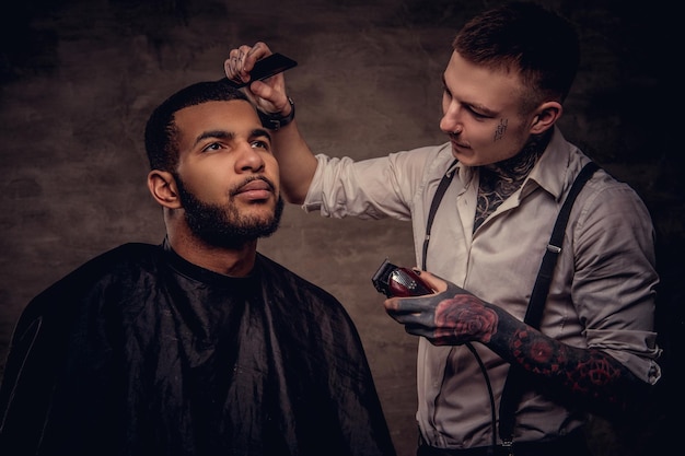 Um cara de hipster afro-americano barbudo cortando o cabelo por um cabeleireiro profissional tatuado à moda antiga faz o corte de cabelo.
