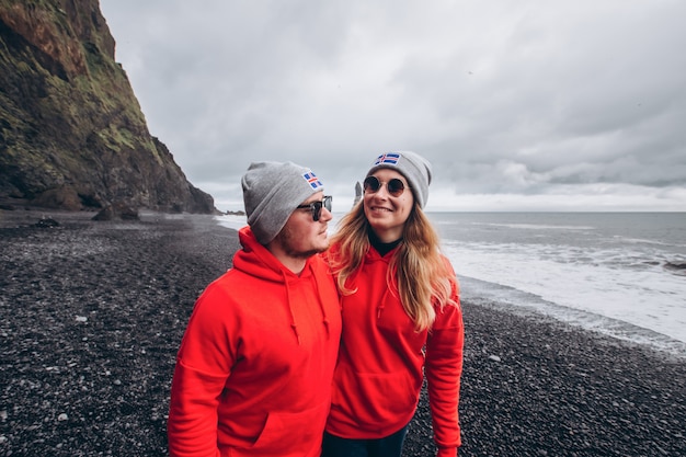 Um cara e uma garota com suéteres vermelhos e chapéus cinza estão se abraçando em uma praia negra, ao lado de Vic