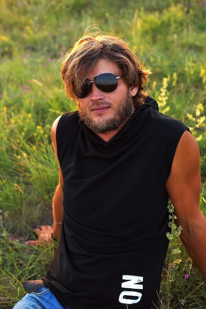 um cara em uma camiseta preta e óculos de sol sentado na grama ao pôr do sol
