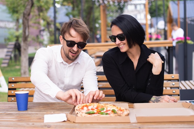 Um cara está sentado com uma garota no terraço de um restaurante e arranca uma pizza