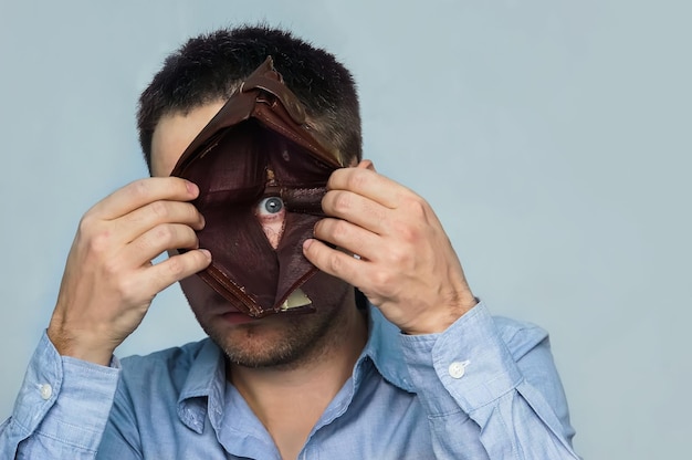 Um cara olha através de um buraco no fundo de uma carteira de couro vazia, mostrando emoções negativas, frustração, desapontamento e tristeza