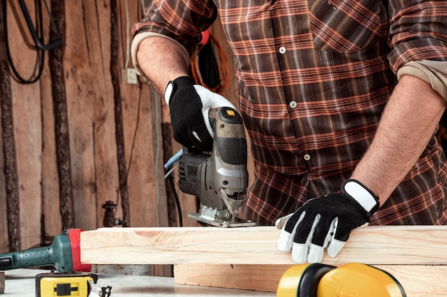 Um carpinteiro de homem corta uma viga de madeira usando um quebra-cabeça elétrico, mãos masculinas com um close de quebra-cabeça elétrico.