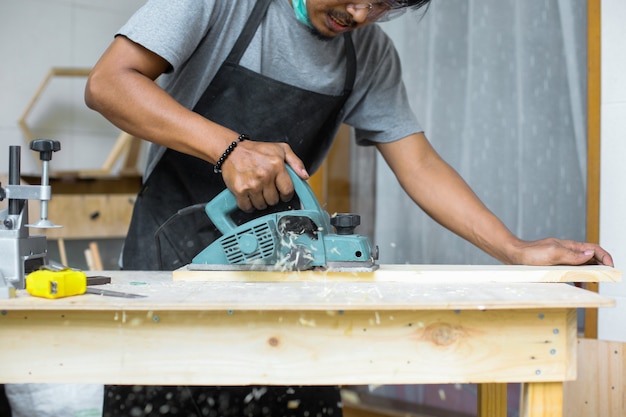 Um carpinteiro trabalhando com uma plaina elétrica em uma prancha de madeira em sua oficina usando equipamento de segurança
