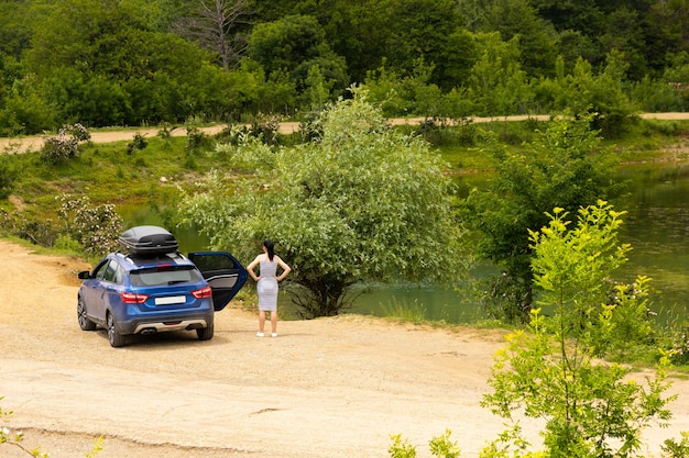 Um carro azul com uma garota com um rack de teto preto está no contexto de um lago de montanha com montanhas e nuvens.