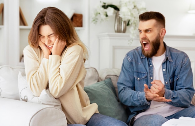 Um casal a discutir, o marido a gritar com a mulher desesperada, sentado no sofá em casa.