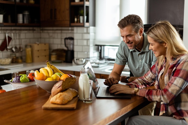 Foto um casal alegre desfruta de um momento alegre em sua cozinha ensolarada trabalhando em um laptop cercado por um pequeno-almoço saudável