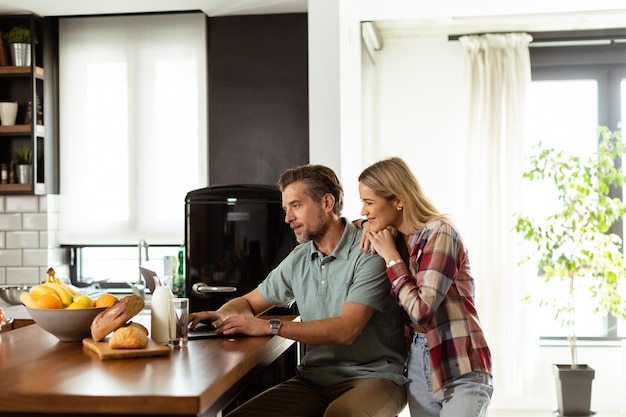 Um casal alegre desfruta de um momento alegre em sua cozinha ensolarada trabalhando em um laptop cercado por um pequeno-almoço saudável