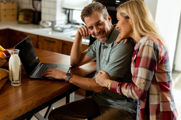 Um casal alegre desfruta de um momento alegre em sua cozinha ensolarada trabalhando em um laptop cercado por um pequeno-almoço saudável