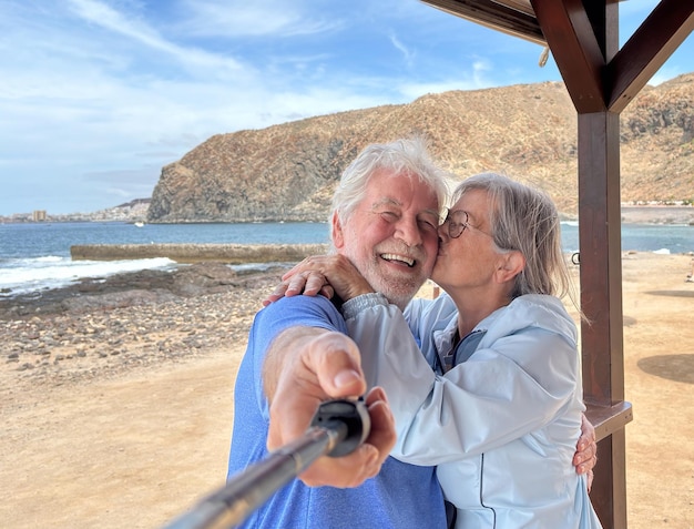 Um casal alegre e sorridente abraçando-se e beijando-se na praia tira fotos com um bastão de selfie