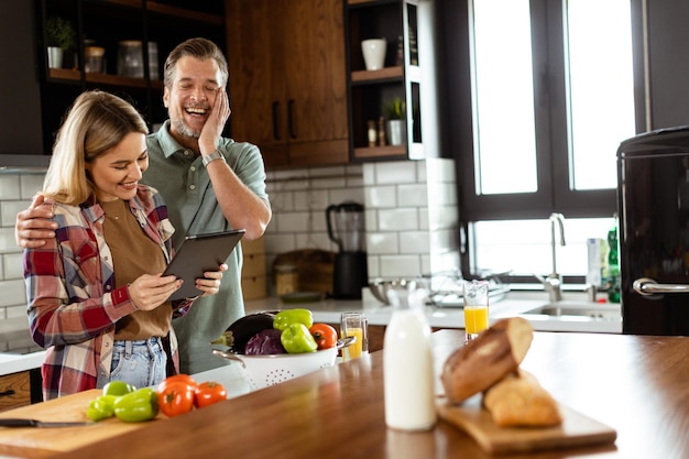 Foto um casal alegre está em uma cozinha bem iluminada absorvido em um tablet digital entre ingredientes frescos