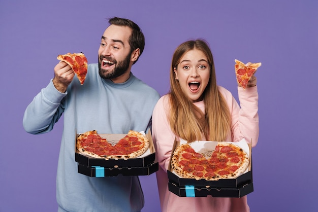 Foto um casal amoroso emocional positivo, amigos isolados sobre a parede roxa, comem pizza.