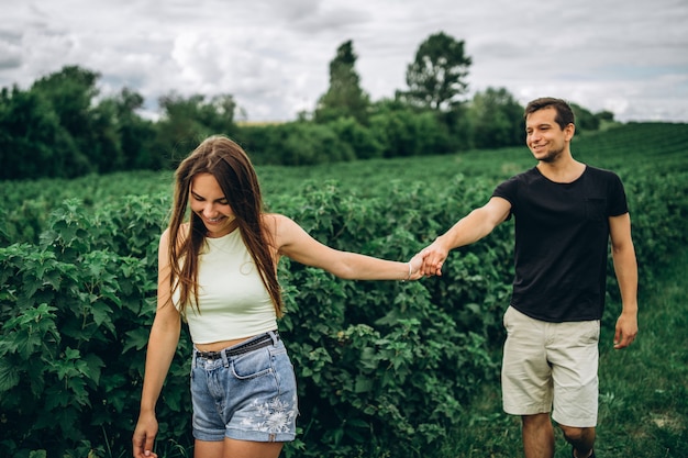 Um casal amoroso terno andando em um campo de groselha. Uma mulher sorridente com cabelos longos leva um homem, segurando sua mão