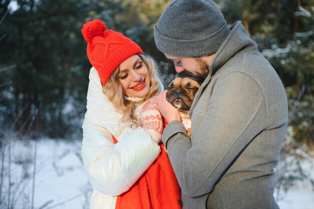 Um casal apaixonado anda pelas mãos no fundo de uma floresta de inverno nevado. Um homem está segurando um cachorro. Uma garota com uma jaqueta branca e um chapéu vermelho.