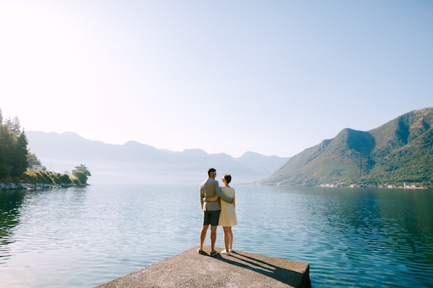 Um casal apaixonado está abraçado em um píer na baía de kotor, perto de perast, tendo como pano de fundo