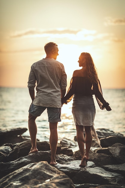 Um casal apaixonado está se divertindo e abraçando na praia vazia do mar ao pôr do sol. Eles estão olhando um ao outro e sorrindo alegremente.