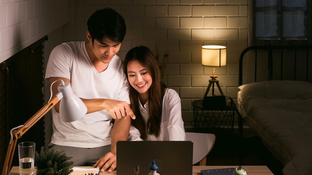 Foto um casal asiático a trabalhar até tarde em casa com um portátil na mesa no quarto escuro.