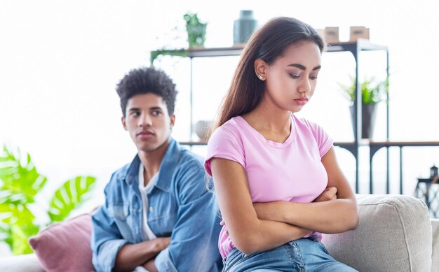 Um casal de adolescentes pretos senta-se num sofá na sala de estar. Ambos estão a olhar para longe um do outro.
