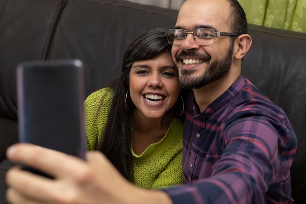 Um casal de amigos latino-americanos sentados no sofá em casa tirando um autorretrato de si mesmos