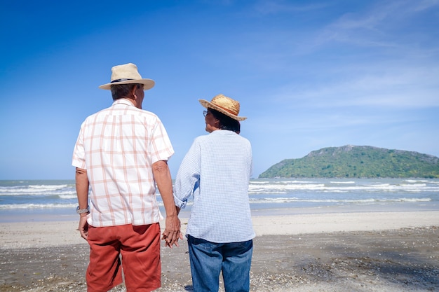 Um casal de idosos asiáticos fica de mãos dadas na praia. olhar para o lindo mar pela manhã, juntos. conceito de viagens para viver feliz na idade da aposentadoria. copie o espaço