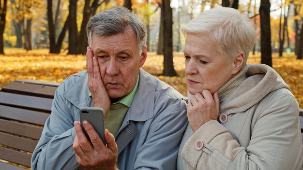 Um casal de idosos chateados senta-se num banco no parque de outono e lê más notícias no smartphone preocupado.