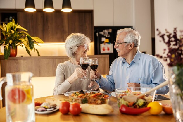 Um casal de idosos feliz está fazendo um brinde com vinho vermelho na mesa de almoço em casa